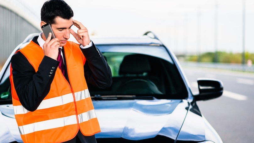A man stands in front of a parked vehicle with a cellphone to his ear while wearing an orange safety vest.