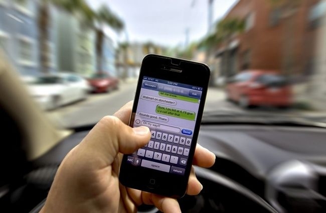 A stock photo of someone texting on an iPhone while driving in front of a blurred background.