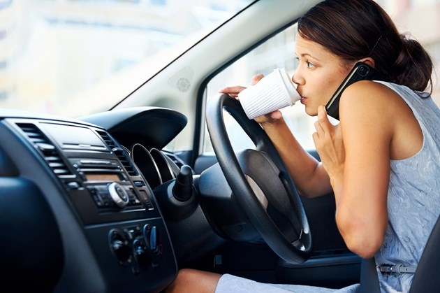 A woman drinks coffee and talks on her phone while attempting to drive in a Shutterstock image.