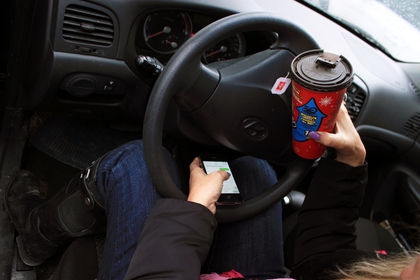 A woman holds a Tim Hortons tea and a cellphone behind the wheel of a vehicle.